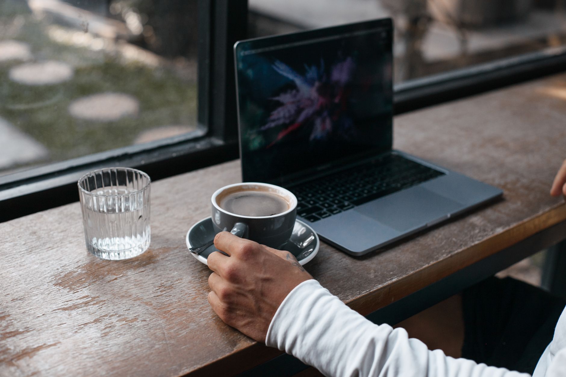 Main tenant une tasse de café à côté d'un ordinateur portable sur une table en bois, près d'une fenêtre.